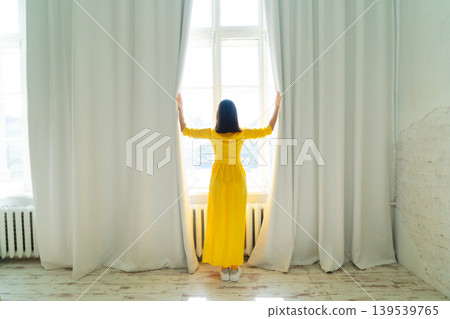 A Cheerful Woman Dressed in a Vibrant Yellow Dress is Posing Gracefully and Happily Near the Bright Window 139539765
