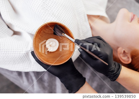 Female beauty therapist applying facial treatment with brush, holding bowl of cream, wearing black gloves, in a serene spa environment, showcasing skincare process and relaxation Female beauty therapist applying facial treatment with brush, holding bowl of cream, wearing black gloves, in a serene spa environment, showcasing skincare process and relaxation 139539850