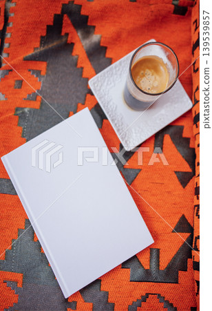 Blank notebook placed on vibrant patterned fabric beside a cup of coffee on a white plate, creating a cozy atmosphere for writing or creative inspiration Blank notebook placed on vibrant patterned fabric beside a cup of coffee on a white plate, creating a cozy atmosphere for writing or creative inspiration 139539857