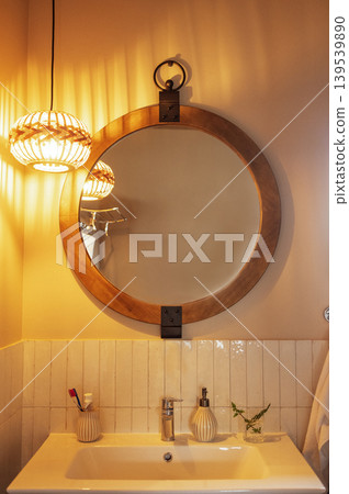 A round mirror in a wooden frame and a lamp with a wicker shade over the washbasin. A close-up of a part of the bathroom interior. A round mirror in a wooden frame and a lamp with a wicker shade over the washbasin. A close-up of a part of the bathroom interior. 139539890