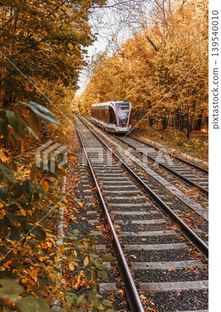 Tram moves along railway tracks through autumn forest corridor with fallen leaves, trees, rails, transport infrastructure. Autumn Travel Route, tram travel, mobility, commuting, tourism, transit route Tram moves along railway tracks through autumn forest corridor with fallen leaves, trees, rails, transport infrastructure. Autumn Travel Route, tram travel, mobility, commuting, tourism, transit route 139540010