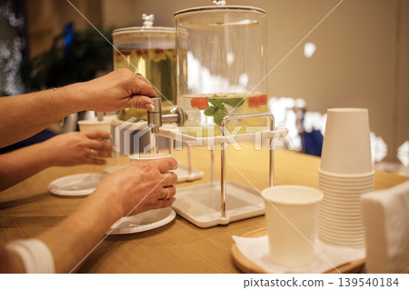 Hands of individuals pouring infused water from beverage dispensers into cups on a wooden table with disposable cups and a blurred background Hands of individuals pouring infused water from beverage dispensers into cups on a wooden table with disposable cups and a blurred background 139540184