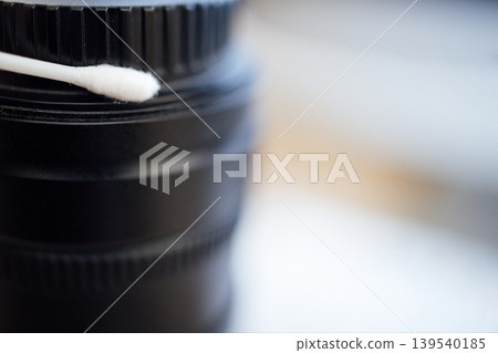 Close-up of a black camera lens with a cotton swab resting on top, showcasing detailed texture and focus on the lens surface and surrounding area Close-up of a black camera lens with a cotton swab resting on top, showcasing detailed texture and focus on the lens surface and surrounding area 139540185