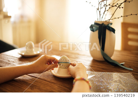 Female hands decorating a white Easter egg with a jeweled band on a wooden table, with a vase and decorative branches in the background Female hands decorating a white Easter egg with a jeweled band on a wooden table, with a vase and decorative branches in the background 139540219