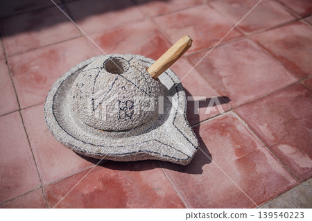 Traditional stone mortar and pestle with wooden handle resting on a tiled surface, showcasing textured details and natural materials in a rustic setting Traditional stone mortar and pestle with wooden handle resting on a tiled surface, showcasing textured details and natural materials in a rustic setting 139540223