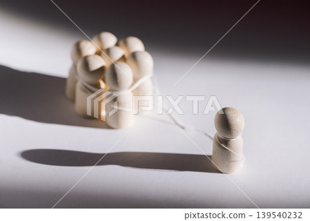 Wooden figurines representing social dynamics, one isolated figure stands apart from a group of connected figures tied with string on a white background 139540232