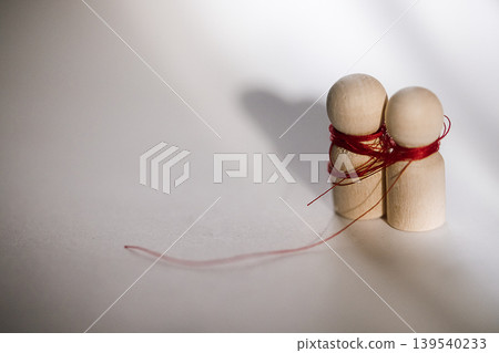 Two wooden figurines wrapped in red thread, positioned closely together on a light background, casting soft shadows that enhance their shapes and textures 139540233