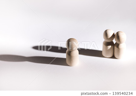 Wooden figurines representing two people standing together and one person standing alone on a white background, casting soft shadows Wooden figurines representing two people standing together and one person standing alone on a white background, casting soft shadows 139540234