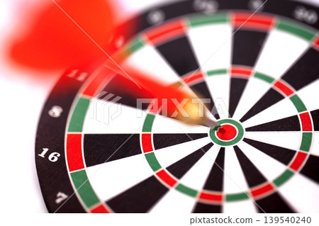 Red dart hitting the bullseye on a dartboard with black and white segments, green and red rings, and numbers visible around the edge of the board Red dart hitting the bullseye on a dartboard with black and white segments, green and red rings, and numbers visible around the edge of the board 139540240
