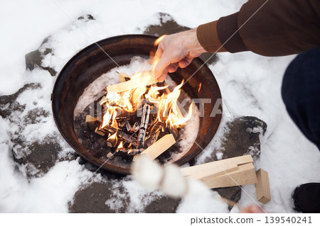 Person adding wood to a fire pit surrounded by snow, with flames visible and a skewer in hand for roasting marshmallows outdoors in a winter setting 139540241
