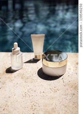 Skincare products including a dropper bottle, cream jar, and tube placed on a stone surface beside a swimming pool with shimmering water in the background 139540250