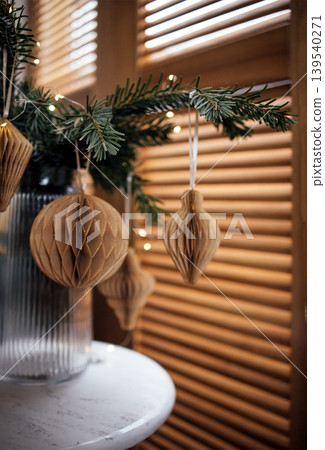 Decorative paper ornaments hanging from a green pine branch, illuminated by soft lights, creating a warm and festive atmosphere for holiday celebrations 139540271