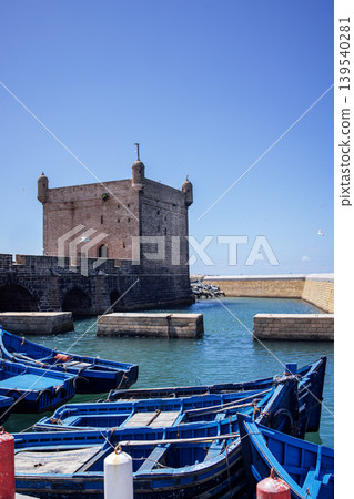 Blue fishing boats are moored in a tranquil harbor, with a historic stone tower standing majestically against a clear blue sky, creating a picturesque coastal scene Blue fishing boats are moored in a tranquil harbor, with a historic stone tower standing majestically against a clear blue sky, creating a picturesque coastal scene 139540281