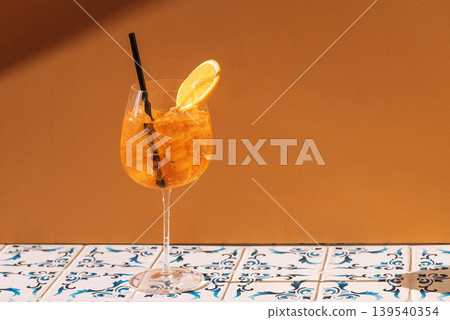 Close-up of a glass with aperol and a piece of orange. A cup of cool, delicious sour orange drink with ice cubes. Citrus alcoholic cocktail and straw on a patterned tile table. Close-up of a glass with aperol and a piece of orange. A cup of cool, delicious sour orange drink with ice cubes. Citrus alcoholic cocktail and straw on a patterned tile table. 139540354