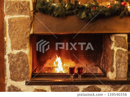 Two glasses with mulled wine are near the fireplace. Christmas decorations and cups with a spicy hot drink. Open fire and stone walls. New Year Eve. Two glasses with mulled wine are near the fireplace. Christmas decorations and cups with a spicy hot drink. Open fire and stone walls. New Year Eve. 139540356