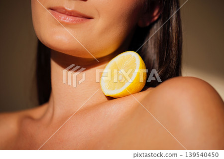 Female model with long dark hair poses with a lemon slice resting on her shoulder, showcasing a close-up view of her skin and the fruit in soft lighting Female model with long dark hair poses with a lemon slice resting on her shoulder, showcasing a close-up view of her skin and the fruit in soft lighting 139540450
