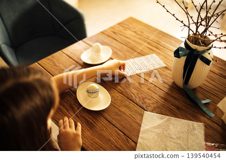Child's hand reaching for decorative Easter egg on wooden table with patterned napkin and vase of twigs, cozy indoor setting with soft lighting Child's hand reaching for decorative Easter egg on wooden table with patterned napkin and vase of twigs, cozy indoor setting with soft lighting 139540454