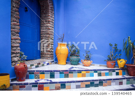 Colorful ceramic pots and plants on tiled steps leading to a blue door in a vibrant courtyard, showcasing traditional Moroccan architecture and design elements 139540465