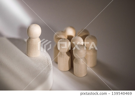 Wooden figurines representing a group dynamic, one figure stands apart on a circular base while others are clustered together in a soft light setting 139540469