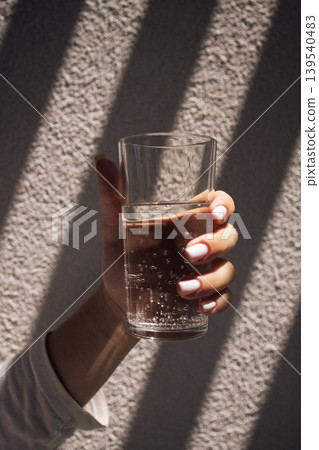 Hand holding a clear glass of sparkling water with bubbles, sunlight casting shadows on a textured wall in the background 139540483