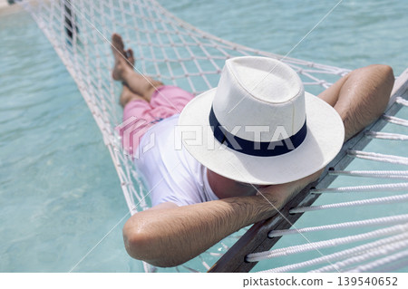 A young man in summer clothes and a hat is lying in a hammock in clear sea water A young man in summer clothes and a hat is lying in a hammock in clear sea water 139540652