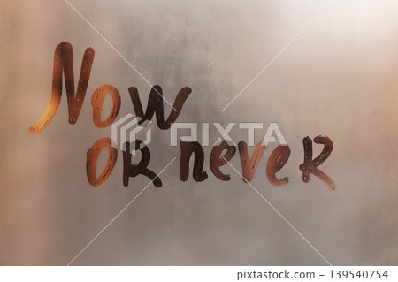 Handwritten message lettering Now or never on misted glass on sunset window with raindrops 139540754