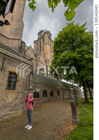 Dordrecht, Zuid-Holland provincie, Netherlands - 09.06.2025: Tourist in pink jacket looking through binoculars at the historic Great Church Grote Kerk in Dordrecht, Netherlands, under a cloudy sky. Dordrecht, Zuid-Holland provincie, Netherlands - 09.06.2025: Tourist in pink jacket looking through binoculars at the historic Great Church Grote Kerk in Dordrecht, Netherlands, under a cloudy sky. 139540843