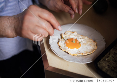 Close-up of hands seasoning a fried egg on a white plate, with a wooden countertop and a baking dish in the background, showcasing culinary preparation Close-up of hands seasoning a fried egg on a white plate, with a wooden countertop and a baking dish in the background, showcasing culinary preparation 139540897
