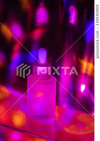 Transparent glass dropper bottle with a white dropper cap illuminated by colorful lights against a blurred vibrant background of pink, purple, and blue hues 139540909
