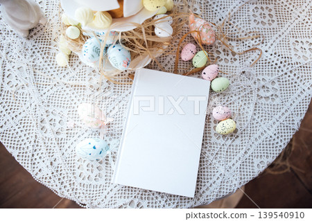 Easter-themed table setting with decorative pastel eggs and a blank notebook on a lace tablecloth, surrounded by festive decorations and a cake stand 139540910
