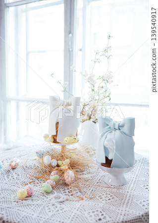Decorative Easter cakes with white and blue bows displayed on white pedestals, surrounded by colorful eggs and spring flowers on a lace tablecloth near a bright window Decorative Easter cakes with white and blue bows displayed on white pedestals, surrounded by colorful eggs and spring flowers on a lace tablecloth near a bright window 139540917