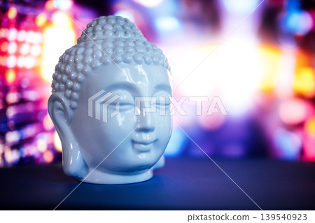 White ceramic Buddha head sculpture with serene expression, placed on a dark surface, surrounded by colorful bokeh lights, creating a tranquil and meditative atmosphere White ceramic Buddha head sculpture with serene expression, placed on a dark surface, surrounded by colorful bokeh lights, creating a tranquil and meditative atmosphere 139540923