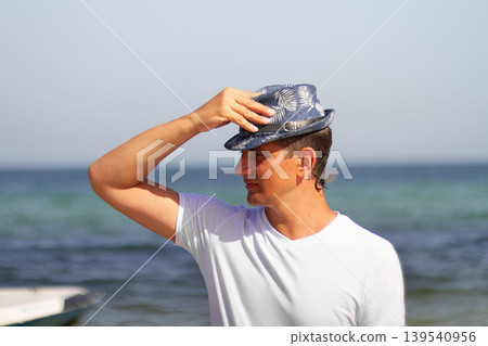 Portrait of middle-aged male tourist of Brazilian appearance in white T-shirt and summer hat against backdrop of sea, copy space 139540956