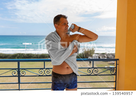 Portrait of sun-tanned middle-aged European male tourist standing in swimming trunks and towel on balcony opposite sea and beach, relax tourism, tour Portrait of sun-tanned middle-aged European male tourist standing in swimming trunks and towel on balcony opposite sea and beach, relax tourism, tour 139540957
