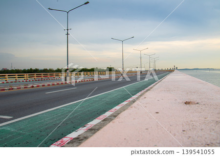 Long perspective of a quiet seaside roadway with evenly spaced streetlights, painted lane markings, and calm water beside the embankment under overcast evening light. Long perspective of a quiet seaside roadway with evenly spaced streetlights, painted lane markings, and calm water beside the embankment under overcast evening light. 139541055
