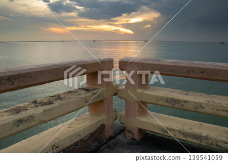 Aged concrete railing overlooks a calm sea under a glowing sunset sky, with soft reflections on the water and layered clouds creating a serene coastal atmosphere. 139541059