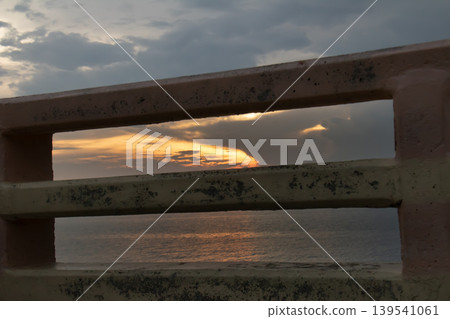 Moody sunset framed by weathered metal railings, with warm golden light reflecting on a calm sea under dramatic clouds. Concept of solitude, barrier, travel, and coastal atmosphere. 139541061
