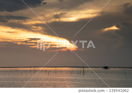 Warm sunset light spreads across shallow coastal waters dotted with fishing stakes and a distant hut. Dramatic clouds create a serene yet powerful seascape, evoking calm, livelihood, and coastal life. Warm sunset light spreads across shallow coastal waters dotted with fishing stakes and a distant hut. Dramatic clouds create a serene yet powerful seascape, evoking calm, livelihood, and coastal life. 139541062