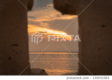 Glowing sunset over shallow sea viewed through corroded metal, revealing rows of fishing stakes and distant horizon. A creative frame adds depth, texture, and a sense of isolation and coastal Glowing sunset over shallow sea viewed through corroded metal, revealing rows of fishing stakes and distant horizon. A creative frame adds depth, texture, and a sense of isolation and coastal 139541063