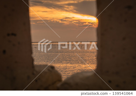 Soft orange sunset seen through aged metal edges, overlooking shallow waters lined with fishing poles. A layered composition blending texture, light, and coastal livelihood in a tranquil atmosphere. Soft orange sunset seen through aged metal edges, overlooking shallow waters lined with fishing poles. A layered composition blending texture, light, and coastal livelihood in a tranquil atmosphere. 139541064
