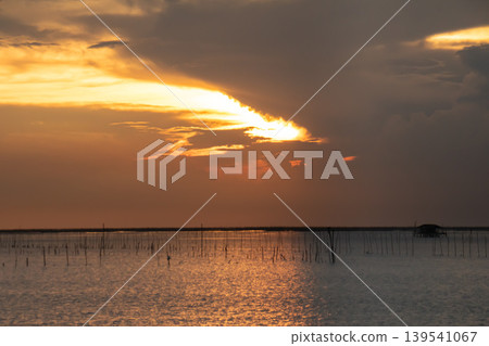 Warm sunset light breaks through clouds over a tranquil sea. Wooden stakes and a small offshore hut create a peaceful coastal scene with shimmering reflections and soft horizon tones. Warm sunset light breaks through clouds over a tranquil sea. Wooden stakes and a small offshore hut create a peaceful coastal scene with shimmering reflections and soft horizon tones. 139541067
