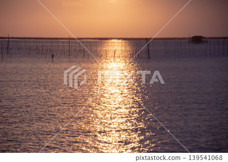 Shimmering path of sunlight stretches across textured water at sunset. Gentle waves and scattered fishing poles create a calm coastal atmosphere with warm pastel sky tones. Shimmering path of sunlight stretches across textured water at sunset. Gentle waves and scattered fishing poles create a calm coastal atmosphere with warm pastel sky tones. 139541068