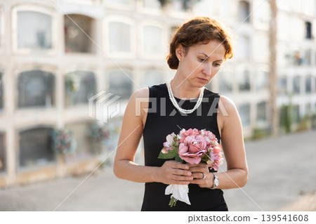 Sad woman in black dress with bouquet of flowers and handkerchief in hands, stands in cemetery 139541608