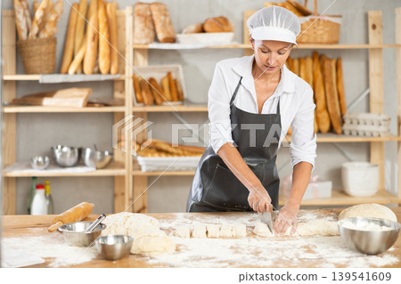 Woman works in bakery as baker, cuts dough into portions, forms pieces of dough to create buns. Woman works in bakery as baker, cuts dough into portions, forms pieces of dough to create buns. 139541609