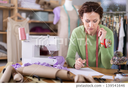 Sad female dressmaker sitting over blank sheet of paper in sewing studio Sad female dressmaker sitting over blank sheet of paper in sewing studio 139541646