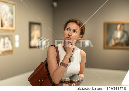 Attractive young woman in casual clothing with phone in hands looking painting in museum 139541718