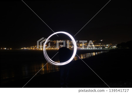 Silhouette of person creating circular light painting on beach at night with city lights Silhouette of person creating circular light painting on beach at night with city lights 139541727