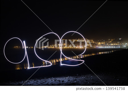 Long exposure light painting of abstract shapes on pebbly beach with city night lights Long exposure light painting of abstract shapes on pebbly beach with city night lights 139541738