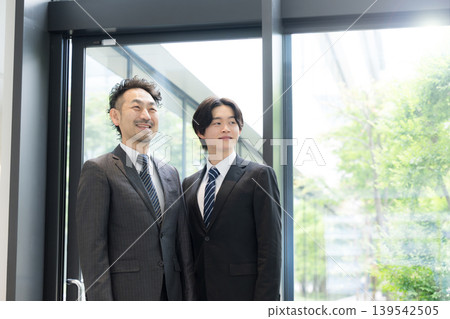 A young businessman and his boss look up from the office window amidst the fresh greenery. A young businessman and his boss look up from the office window amidst the fresh greenery. 139542505