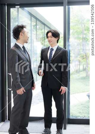 A young businessman and his boss converse by the window in the office surrounded by fresh greenery, discussing business negotiations with clients and their growth. A young businessman and his boss converse by the window in the office surrounded by fresh greenery, discussing business negotiations with clients and their growth. 139542506